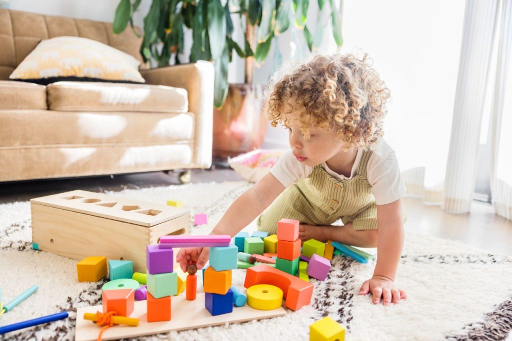 Curly-haired toddler ignoring instructions, refusing to stop and continuing to play with colorful wooden blocks on the living room floor.
