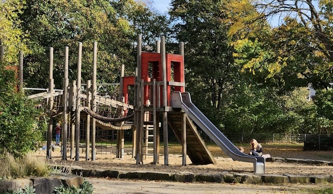 Spielplatz Dormagener Straße – Wooden climbing structure and slide at an outdoor playground in Düsseldorf surrounded by autumn trees
