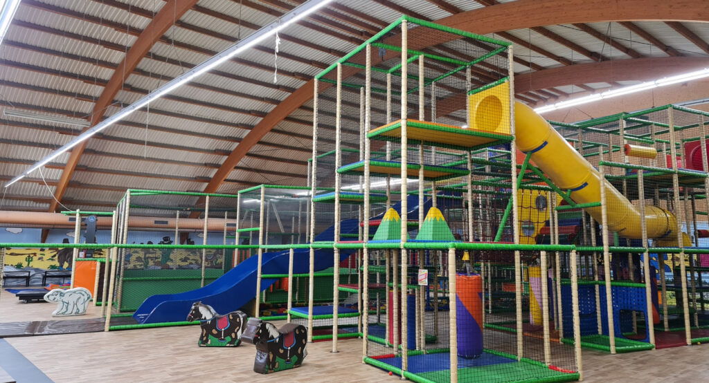 Large indoor play structure with colorful tunnels, slides, climbing nets, and animal-themed ride-on toys under a curved wooden roof