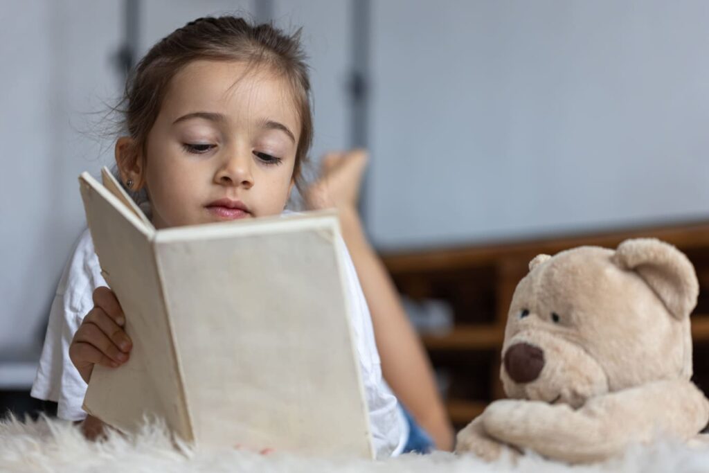Preschool child looking at a book independently next to a teddy bear, exploring stories and developing interest in reading