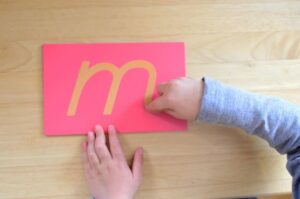 Child tracing Montessori sandpaper letter to build tactile letter recognition and phonemic awareness, a cornerstone of Montessori writing before reading instruction.

