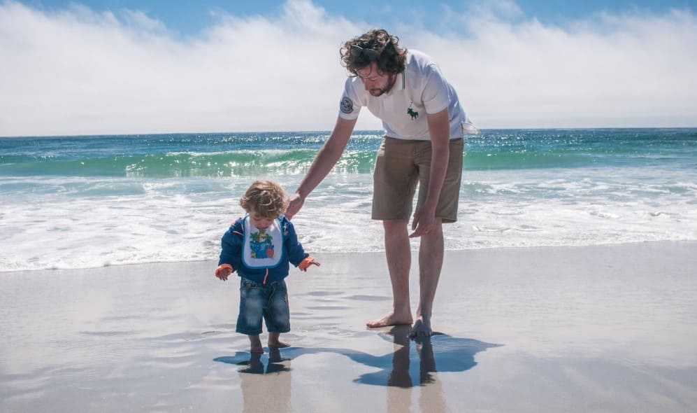 Toddler exploring the beach with a parent’s gentle guidance, demonstrating key principles of Montessori child-led outdoor learning and sensory exploration.

