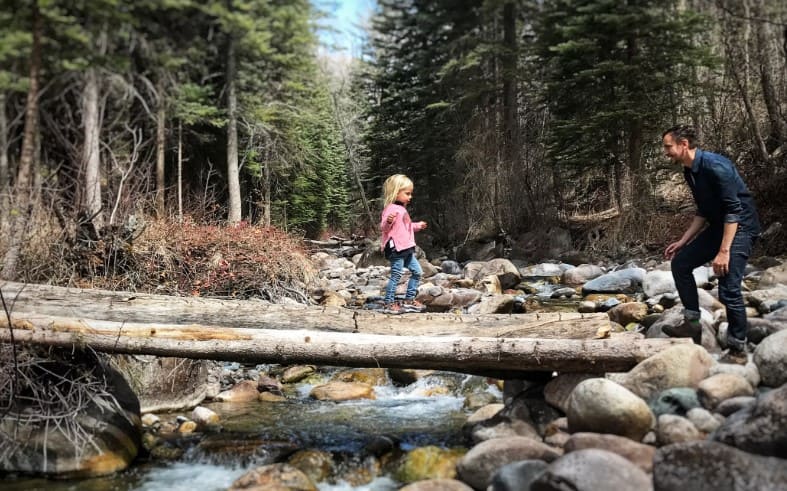 Child walking across a log over a creek with parent nearby, showcasing the benefits of Montessori child-led walks such as independence, confidence, and nature-based discovery.

