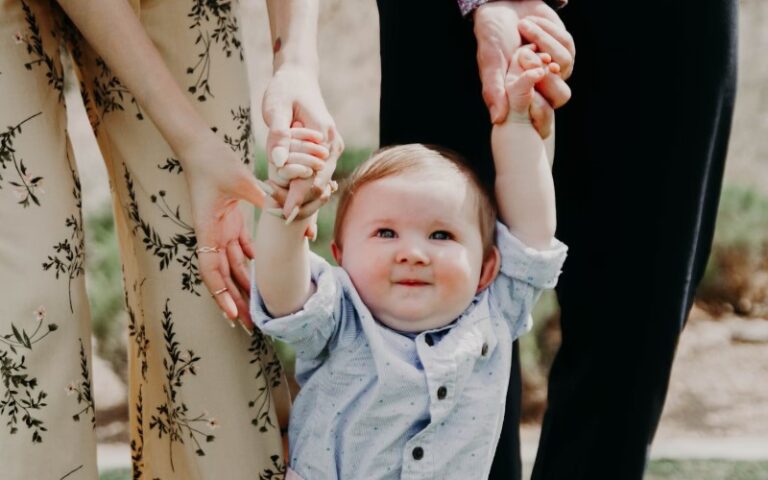 Baby holding hands with adults while walking outdoors, capturing the early stages of a Montessori child-led walk and the development of movement, trust, and exploration.