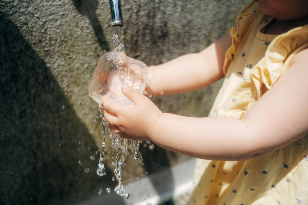 Toddler filling a cup under running water — Montessori practical life activity showing real experience first, which later supports symbolic play and pretend scenarios.