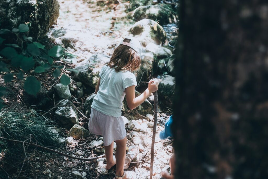 A child navigating uneven forest terrain near a stream, developing balance, concentration, and self-regulation through direct interaction with nature as a teacher.